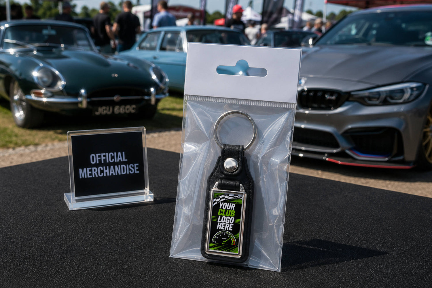 Car Club personalised leather and metal keyring in packing. Displayed on table next to Official Merchandise sign at car  show with BMW and Jaguar in background.